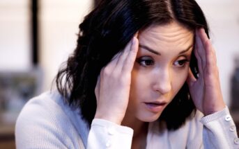 A woman with dark hair holds her hands to her temples, appearing stressed or deep in thought. She is wearing a light-colored sweater and looking slightly downward. The background is softly blurred.