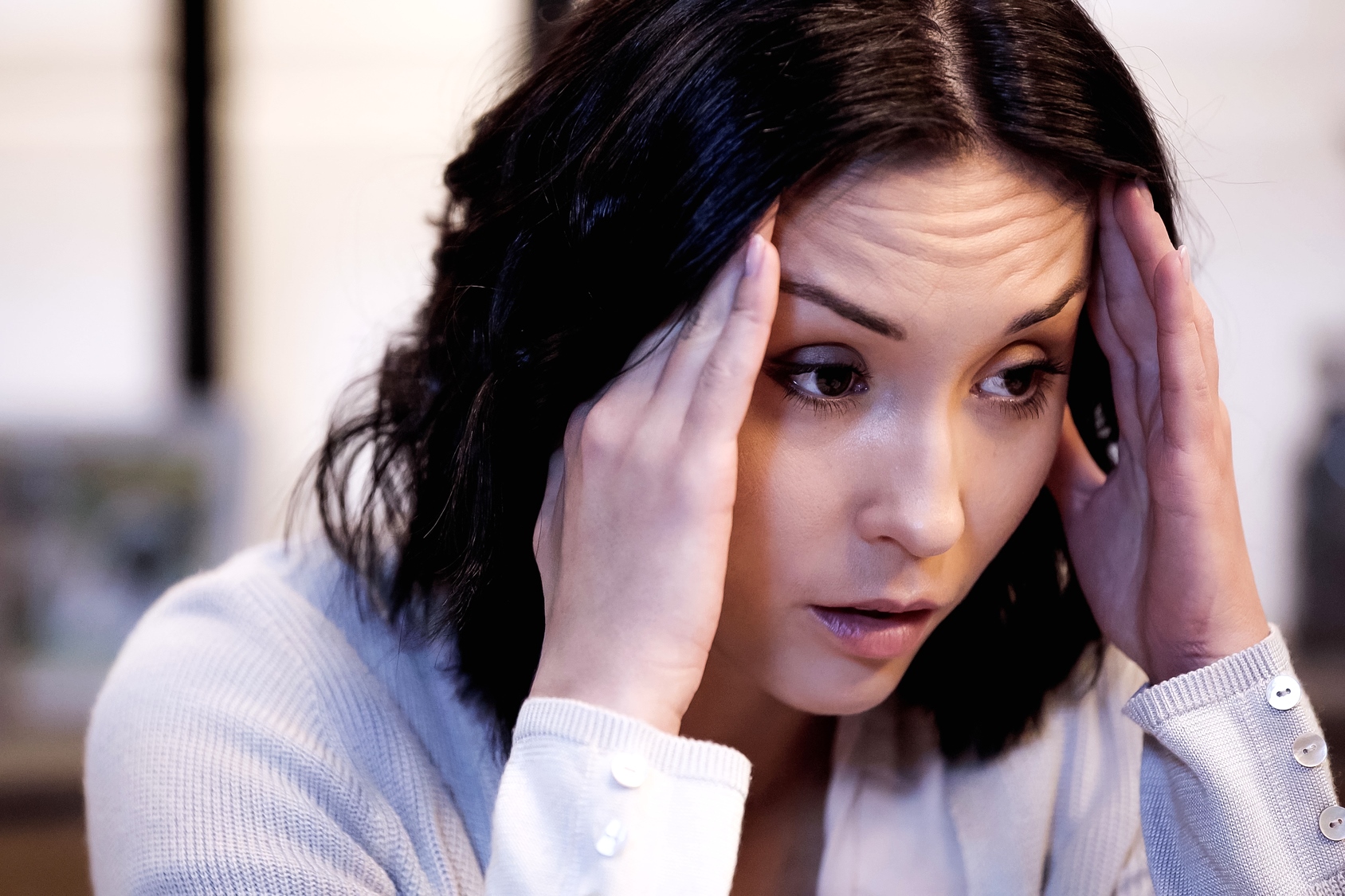 A woman with dark hair holds her hands to her temples, appearing stressed or deep in thought. She is wearing a light-colored sweater and looking slightly downward. The background is softly blurred.