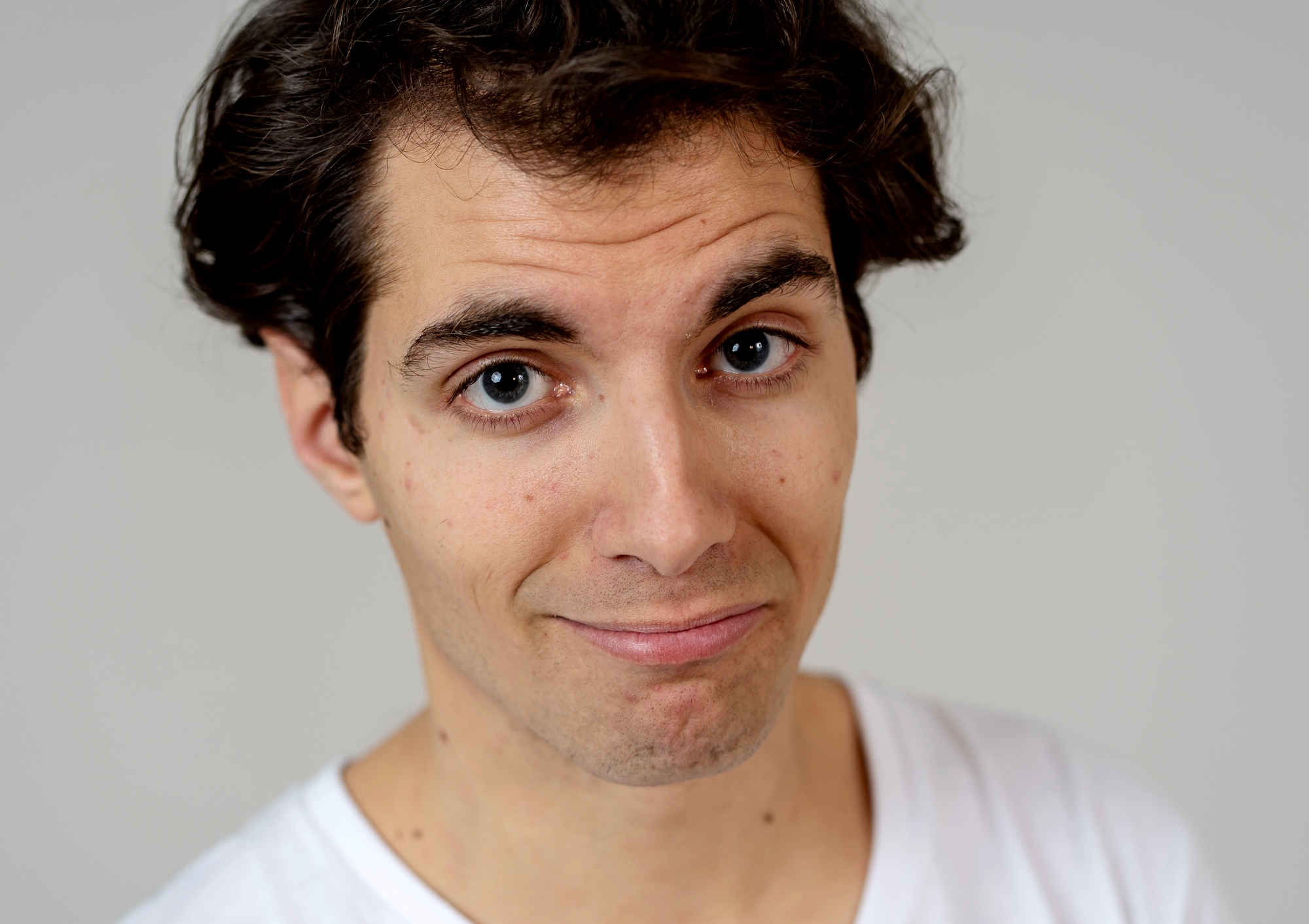 A person with short brown hair wearing a white shirt is slightly smiling while looking at the camera against a plain background.