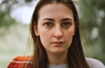 A woman with long brown hair and a neutral expression stands outdoors. She is wearing a gray shirt and a red plaid scarf. The background is blurred with greenery.