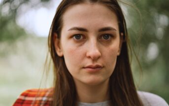 A woman with long brown hair and a neutral expression stands outdoors. She is wearing a gray shirt and a red plaid scarf. The background is blurred with greenery.