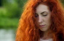 A person with long, vibrant red hair stands outdoors, looking down thoughtfully. The background is blurred with green foliage, suggesting a natural setting. Bright sunlight illuminates the person's face and hair.