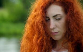 A person with long, vibrant red hair stands outdoors, looking down thoughtfully. The background is blurred with green foliage, suggesting a natural setting. Bright sunlight illuminates the person's face and hair.