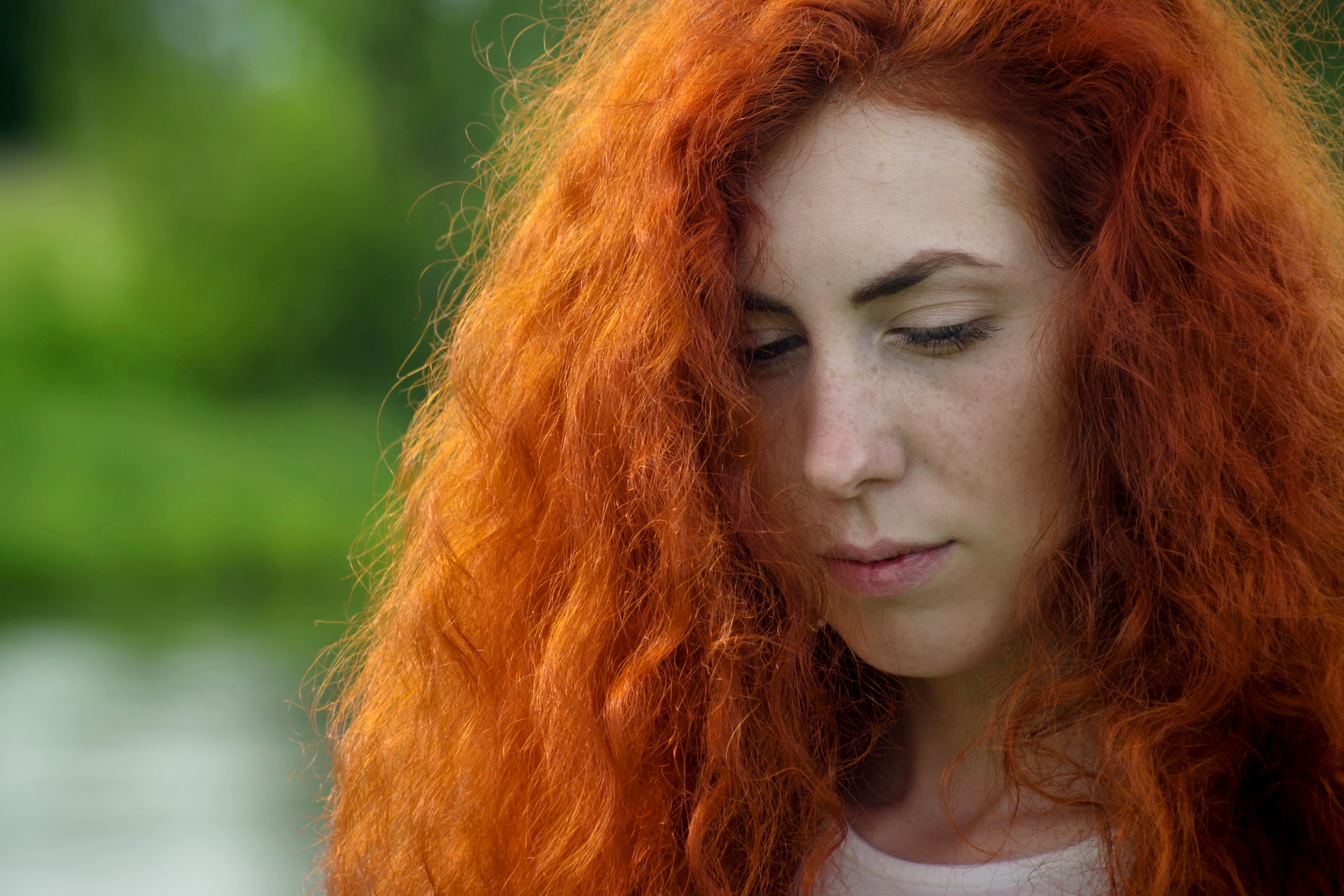 A person with long, vibrant red hair stands outdoors, looking down thoughtfully. The background is blurred with green foliage, suggesting a natural setting. Bright sunlight illuminates the person's face and hair.