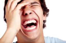 A person laughing energetically with eyes closed and hand on forehead, showing white teeth, set against a white background.