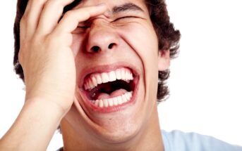 A person laughing energetically with eyes closed and hand on forehead, showing white teeth, set against a white background.