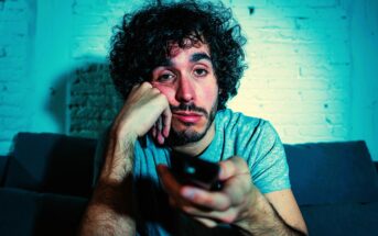 A man with curly hair and a beard sits on a couch, looking bored or tired. He's holding a TV remote in his hand, resting his chin on his other hand, in a dimly lit room with a brick wall background.