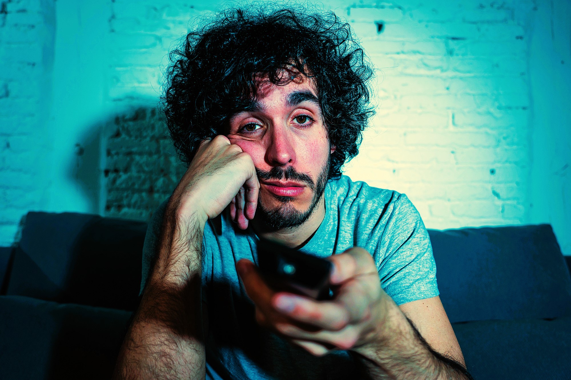 A man with curly hair and a beard sits on a couch, looking bored or tired. He's holding a TV remote in his hand, resting his chin on his other hand, in a dimly lit room with a brick wall background.