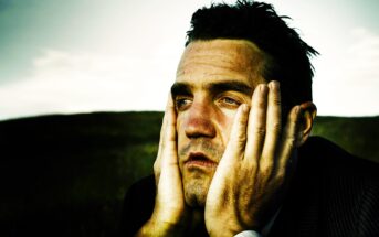 Man with short dark hair wearing a suit appears deep in thought, resting his face in his hands. The background is a blurred field and sky, creating a contemplative mood.