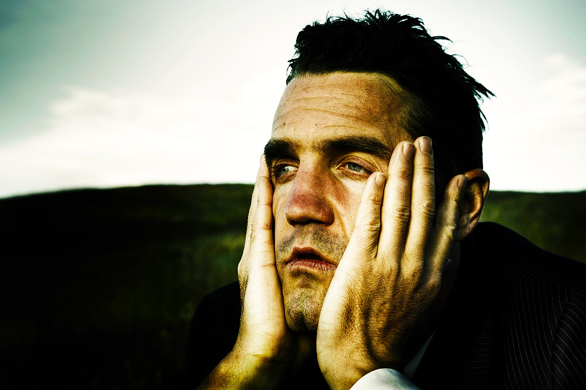 Man with short dark hair wearing a suit appears deep in thought, resting his face in his hands. The background is a blurred field and sky, creating a contemplative mood.