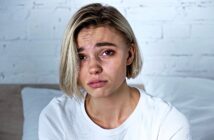 A person with short blonde hair wearing a white shirt appears upset, with slightly furrowed brows and tearful eyes. They are indoors, in front of a white brick wall and sitting on a bed.