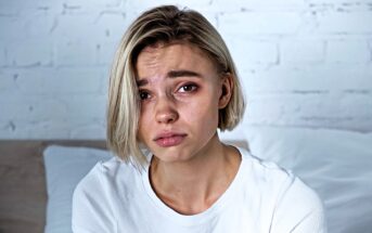 A person with short blonde hair wearing a white shirt appears upset, with slightly furrowed brows and tearful eyes. They are indoors, in front of a white brick wall and sitting on a bed.