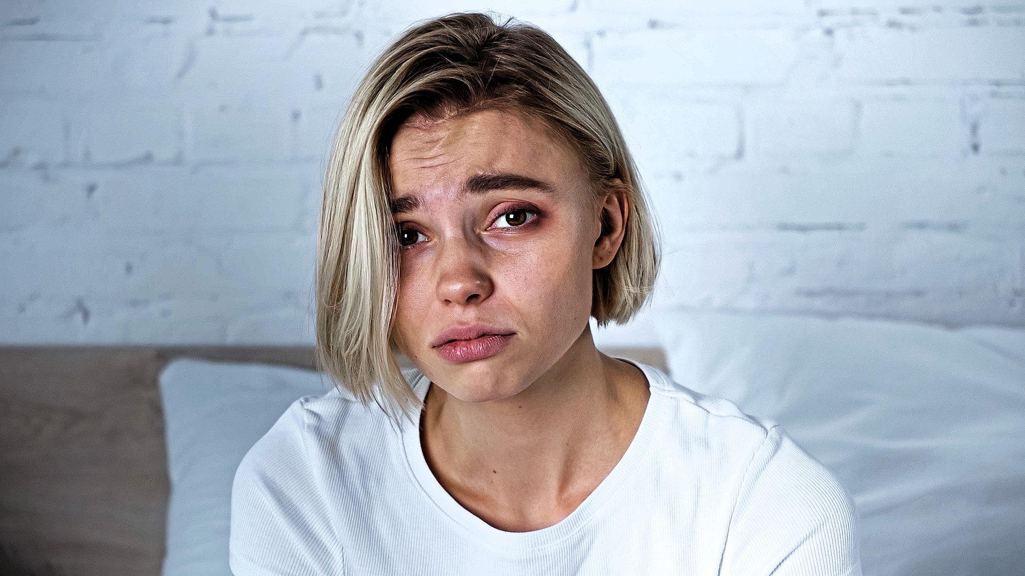 A person with short blonde hair wearing a white shirt appears upset, with slightly furrowed brows and tearful eyes. They are indoors, in front of a white brick wall and sitting on a bed.