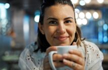 A person smiling warmly and holding a white mug in a cozy, softly lit setting. The background features decorative bokeh lights.