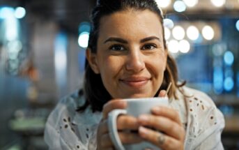 A person smiling warmly and holding a white mug in a cozy, softly lit setting. The background features decorative bokeh lights.