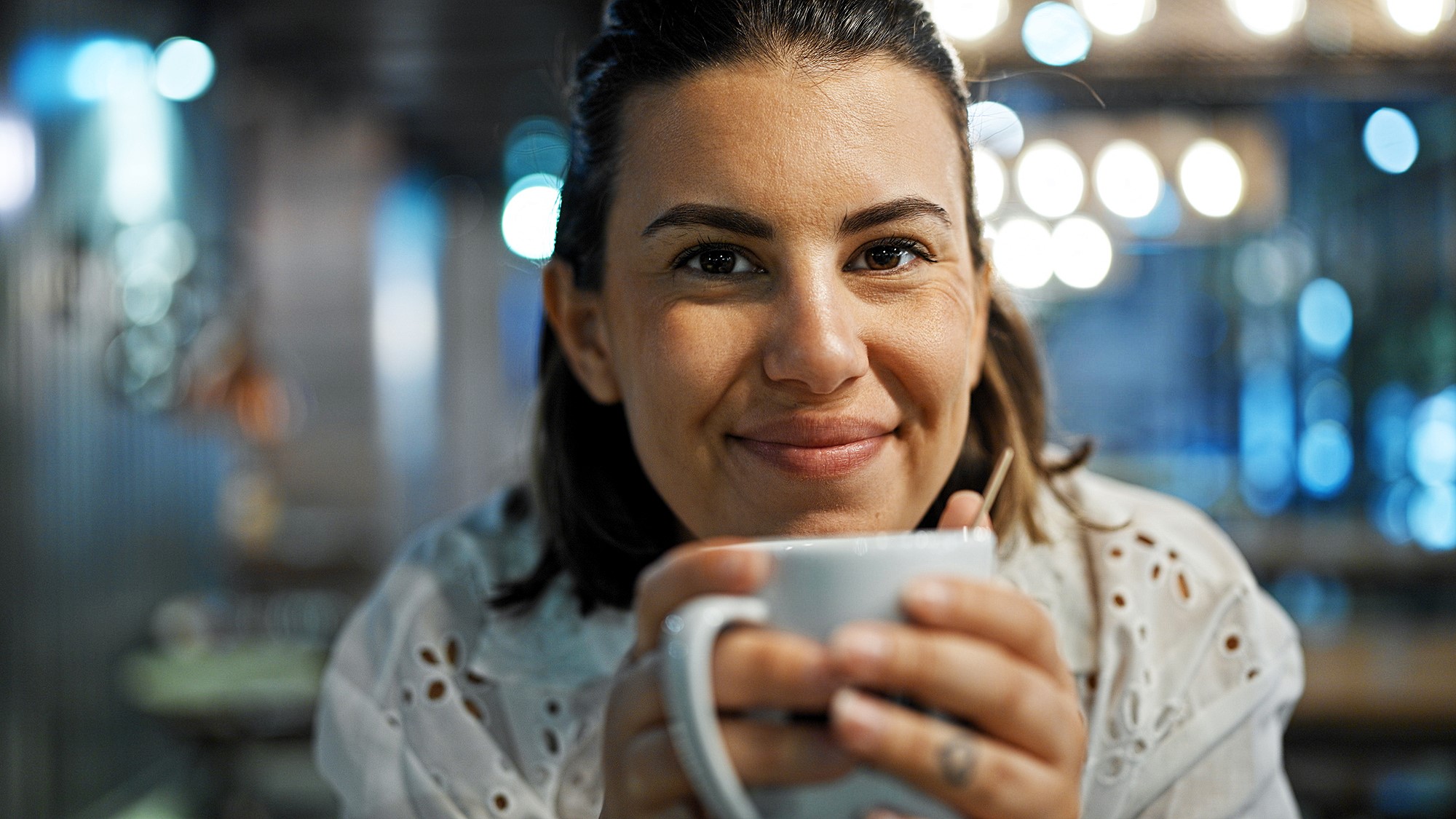 A person smiling warmly and holding a white mug in a cozy, softly lit setting. The background features decorative bokeh lights.