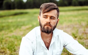 A man with a beard and short hair, wearing a white shirt, sits outdoors in a grassy field. He has a thoughtful expression and looks off into the distance, with a backdrop of trees and cloudy sky.