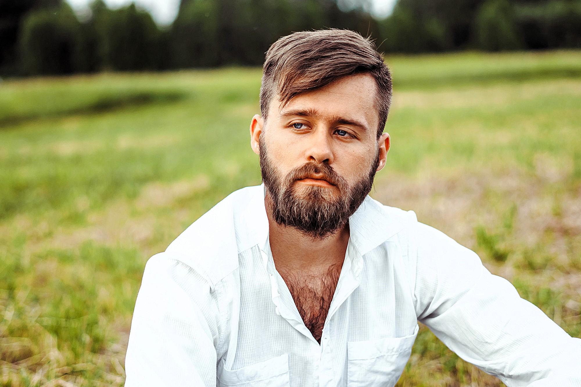 A man with a beard and short hair, wearing a white shirt, sits outdoors in a grassy field. He has a thoughtful expression and looks off into the distance, with a backdrop of trees and cloudy sky.