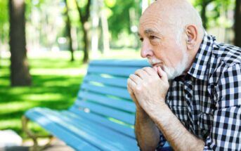 Elderly man with a beard sitting on a blue bench in a park. He rests his chin on his hands, looking contemplative. Trees and greenery are visible in the background.