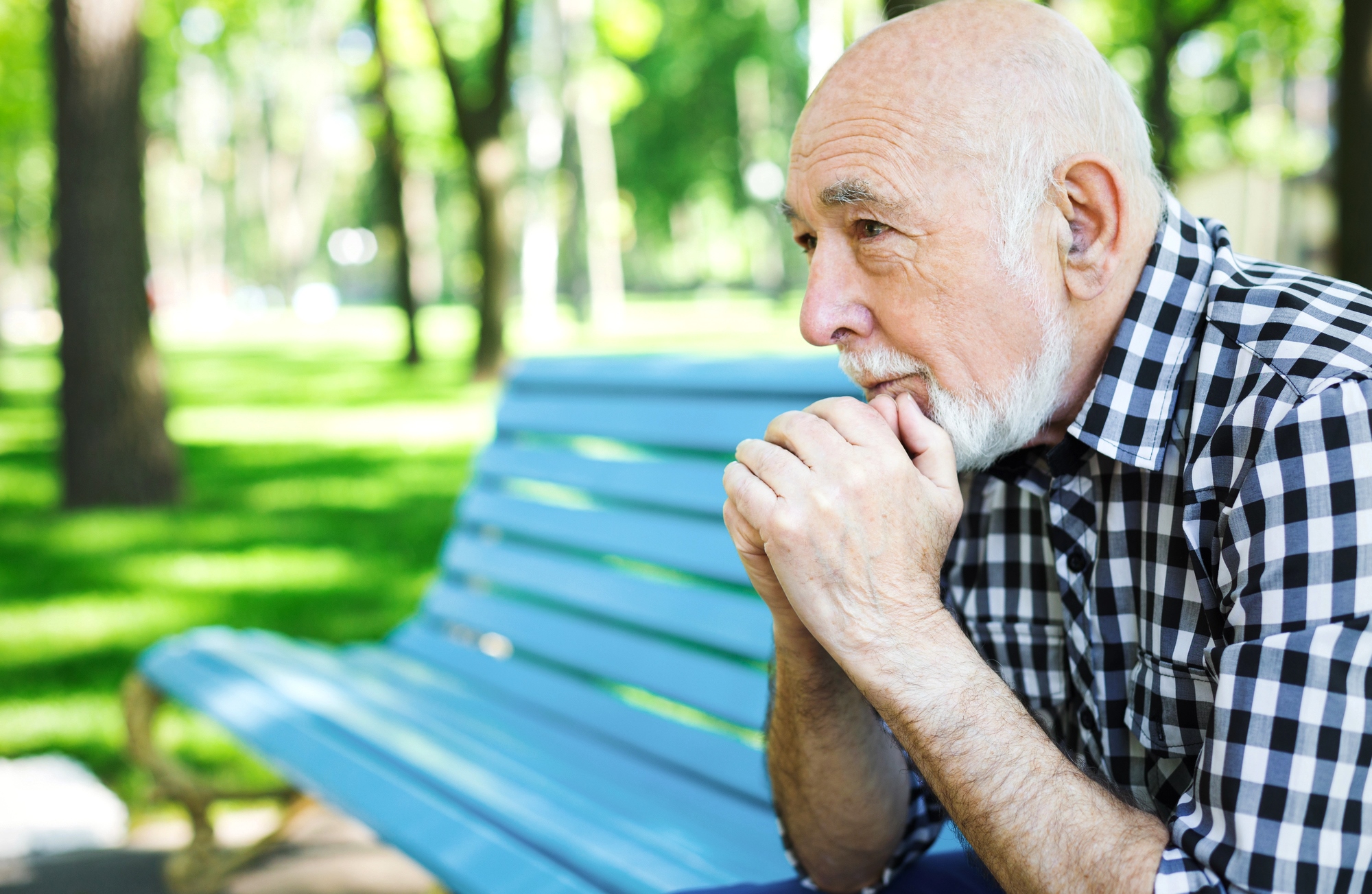 Elderly man with a beard sitting on a blue bench in a park. He rests his chin on his hands, looking contemplative. Trees and greenery are visible in the background.