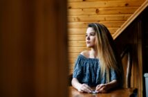 A woman with long hair sits at a wooden table, looking thoughtfully to the side. She is wearing an off-the-shoulder blue patterned top. The background features wooden paneled walls.