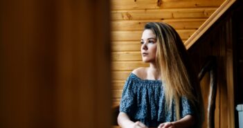 A woman with long hair sits at a wooden table, looking thoughtfully to the side. She is wearing an off-the-shoulder blue patterned top. The background features wooden paneled walls.