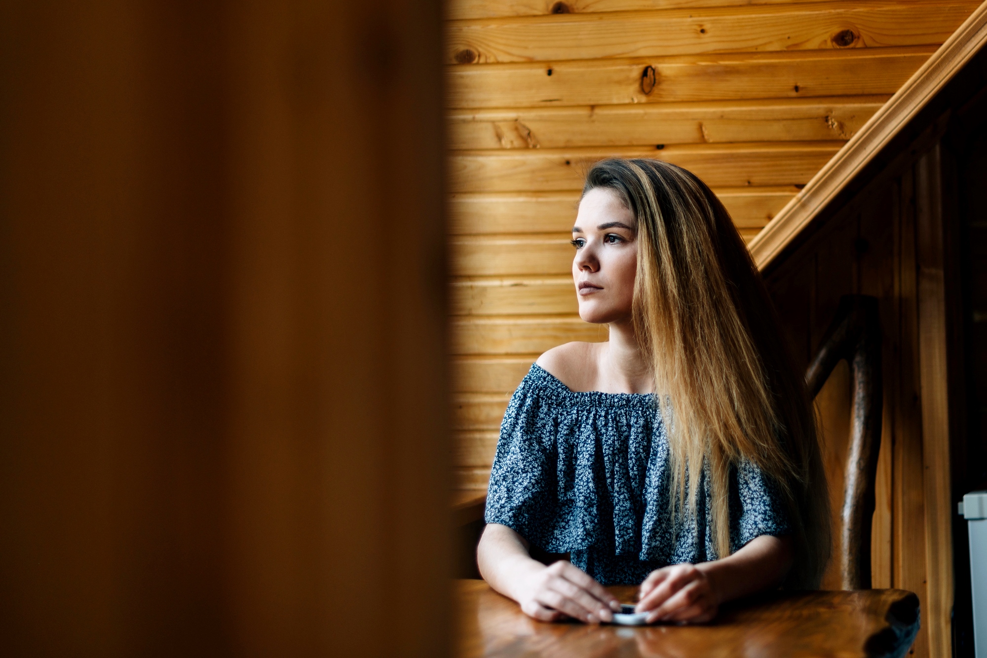 A woman with long hair sits at a wooden table, looking thoughtfully to the side. She is wearing an off-the-shoulder blue patterned top. The background features wooden paneled walls.