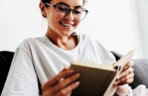 A woman with glasses is smiling while reading a book. She is wearing a white shirt and sitting on a couch. The background is softly blurred, creating a cozy and relaxed atmosphere.