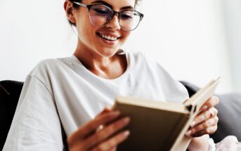 A woman with glasses is smiling while reading a book. She is wearing a white shirt and sitting on a couch. The background is softly blurred, creating a cozy and relaxed atmosphere.