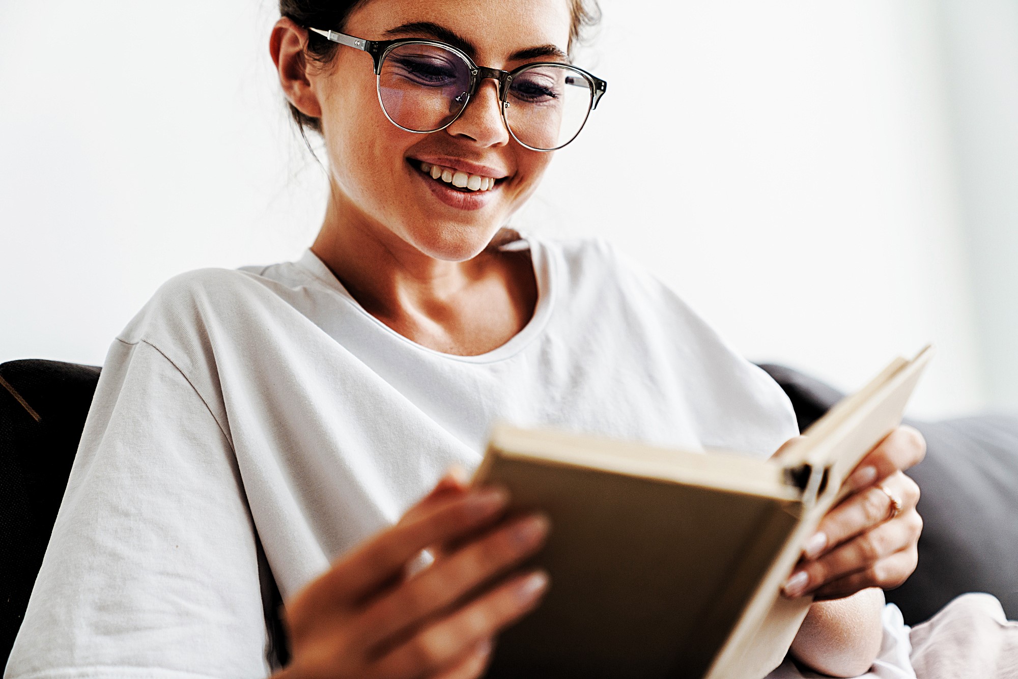 A woman with glasses is smiling while reading a book. She is wearing a white shirt and sitting on a couch. The background is softly blurred, creating a cozy and relaxed atmosphere.