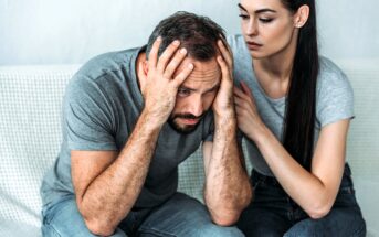 A man sits on a couch with his head in his hands, looking distressed. A woman sits beside him, gently touching his shoulder, offering support. Both are wearing casual gray shirts and jeans.