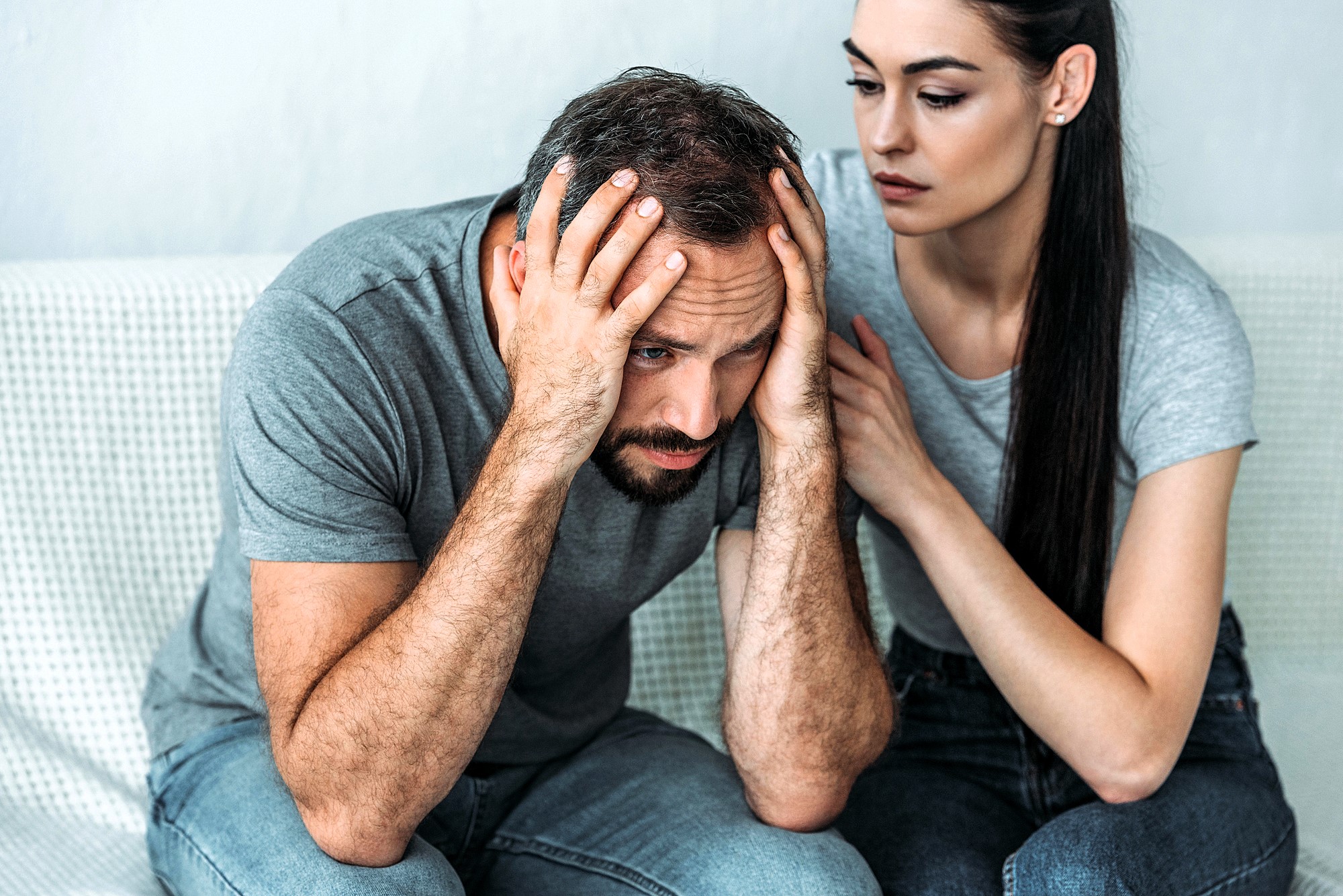 A man sits on a couch with his head in his hands, looking distressed. A woman sits beside him, gently touching his shoulder, offering support. Both are wearing casual gray shirts and jeans.