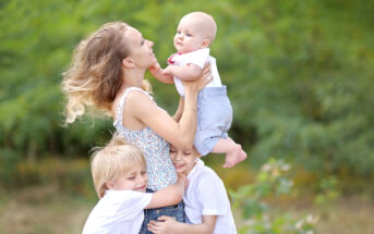 A woman joyfully holds a baby in her arms while two children hug her waist in a park. The group is smiling, surrounded by greenery, creating a cheerful and loving scene.