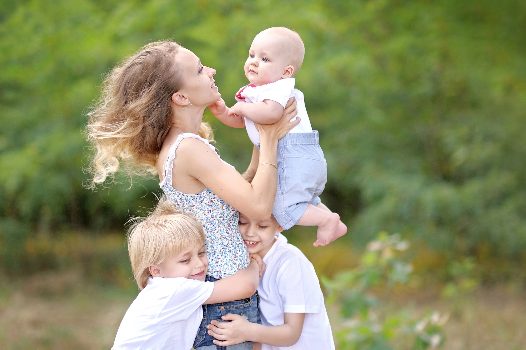 A woman joyfully holds a baby in her arms while two children hug her waist in a park. The group is smiling, surrounded by greenery, creating a cheerful and loving scene.