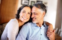 A smiling couple with graying hair embraces warmly. The woman is wearing a light-colored top, and the man is in a blue shirt. The background is softly blurred, suggesting a cozy indoor setting.