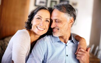 A smiling couple with graying hair embraces warmly. The woman is wearing a light-colored top, and the man is in a blue shirt. The background is softly blurred, suggesting a cozy indoor setting.