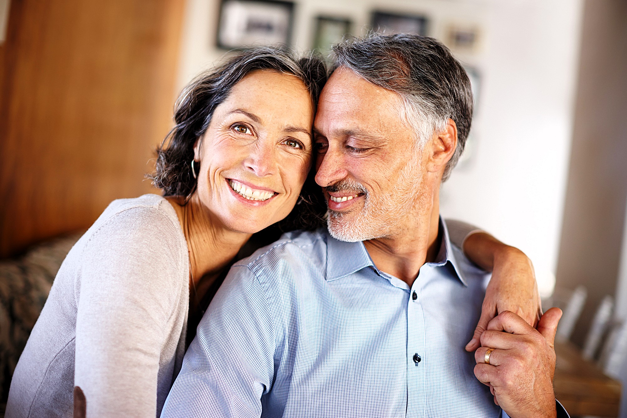 A smiling couple with graying hair embraces warmly. The woman is wearing a light-colored top, and the man is in a blue shirt. The background is softly blurred, suggesting a cozy indoor setting.