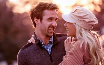 A couple smiling lovingly at each other in a warm, outdoor setting. The man is wearing a dark sweater, and the woman is in a light hat and sweater. The background is softly blurred, suggesting a sunset or sunrise.