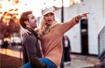 A smiling man in a gray sweater lifts a woman in an orange sweater and beige cap, who points excitedly into the distance. They are outdoors with a soft-focus background of trees and structures, under a warm, golden light.