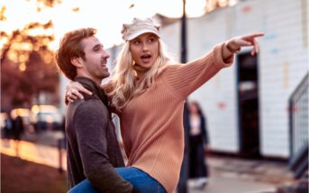 A smiling man in a gray sweater lifts a woman in an orange sweater and beige cap, who points excitedly into the distance. They are outdoors with a soft-focus background of trees and structures, under a warm, golden light.