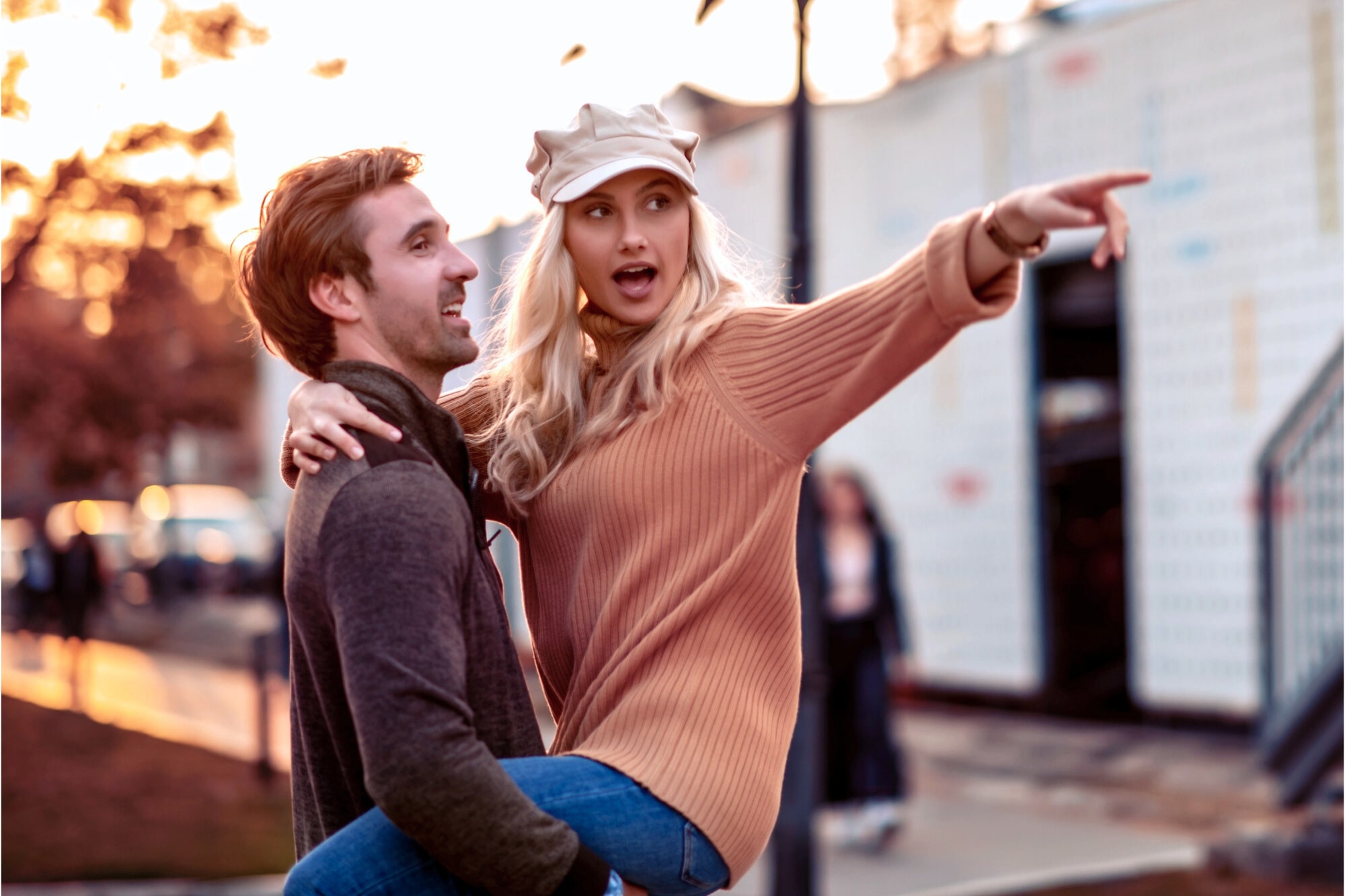 A smiling man in a gray sweater lifts a woman in an orange sweater and beige cap, who points excitedly into the distance. They are outdoors with a soft-focus background of trees and structures, under a warm, golden light.