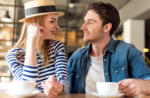 A man and woman sit at a café table with coffee cups. The woman, wearing a striped shirt and straw hat, smiles at the man in a denim jacket. They look at each other warmly, suggesting a pleasant conversation in a cozy atmosphere.