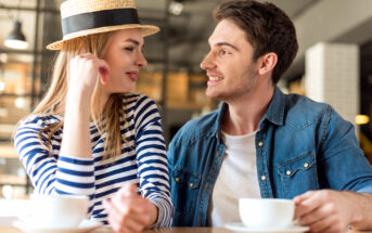 A man and woman sit at a café table with coffee cups. The woman, wearing a striped shirt and straw hat, smiles at the man in a denim jacket. They look at each other warmly, suggesting a pleasant conversation in a cozy atmosphere.