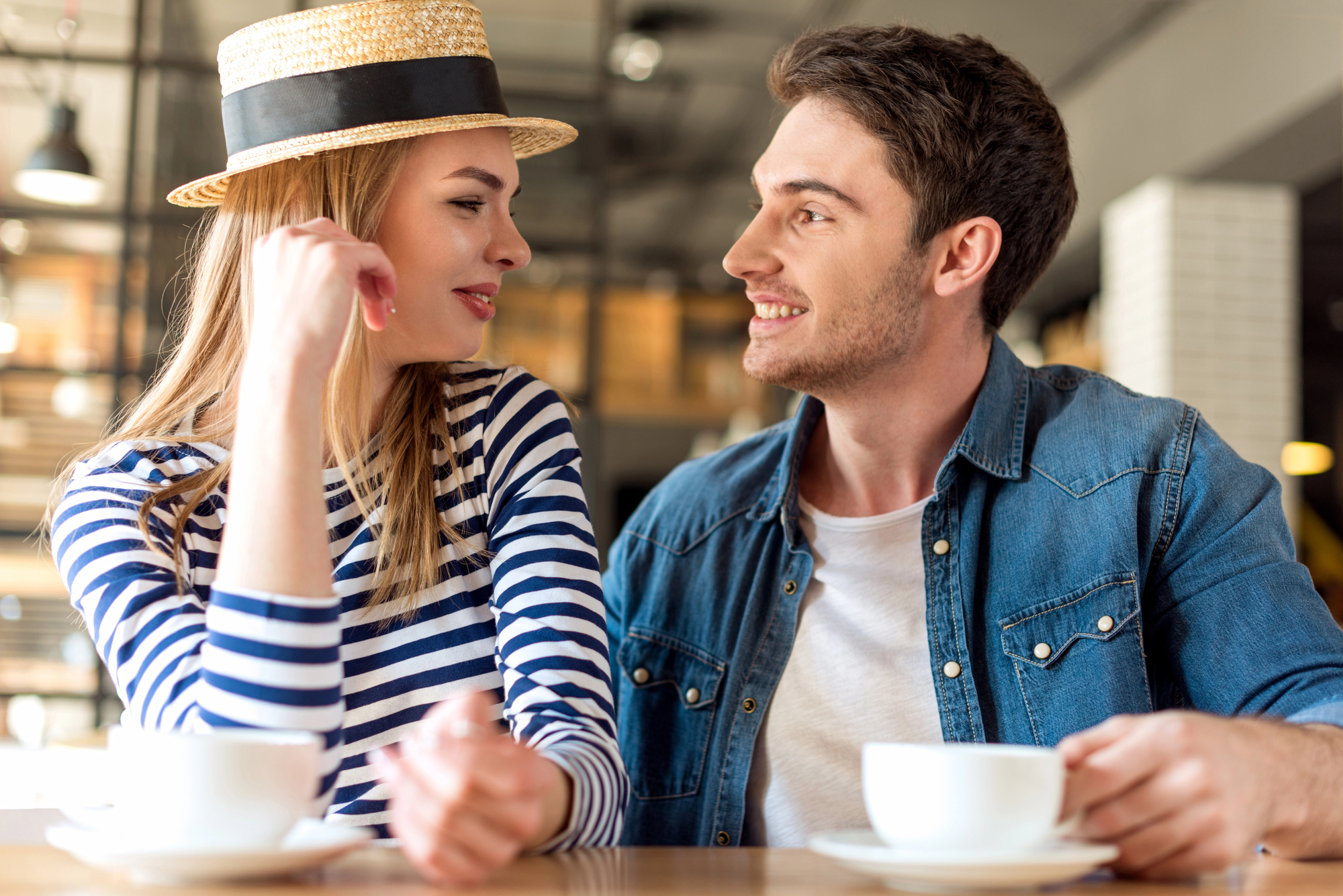 A man and woman sit at a café table with coffee cups. The woman, wearing a striped shirt and straw hat, smiles at the man in a denim jacket. They look at each other warmly, suggesting a pleasant conversation in a cozy atmosphere.