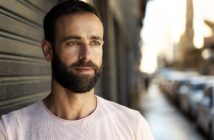 A man with a beard and short hair is leaning against a wall on a city street. He is wearing a light pink T-shirt and looking thoughtfully into the distance. The background is blurred, with cars and buildings in soft focus.