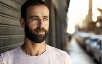 A man with a beard and short hair is leaning against a wall on a city street. He is wearing a light pink T-shirt and looking thoughtfully into the distance. The background is blurred, with cars and buildings in soft focus.