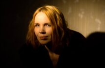 A woman with shoulder-length blonde hair is partially illuminated against a dark background. She is wearing a black top and has a neutral expression. The lighting creates a dramatic effect with shadows on one side of her face.