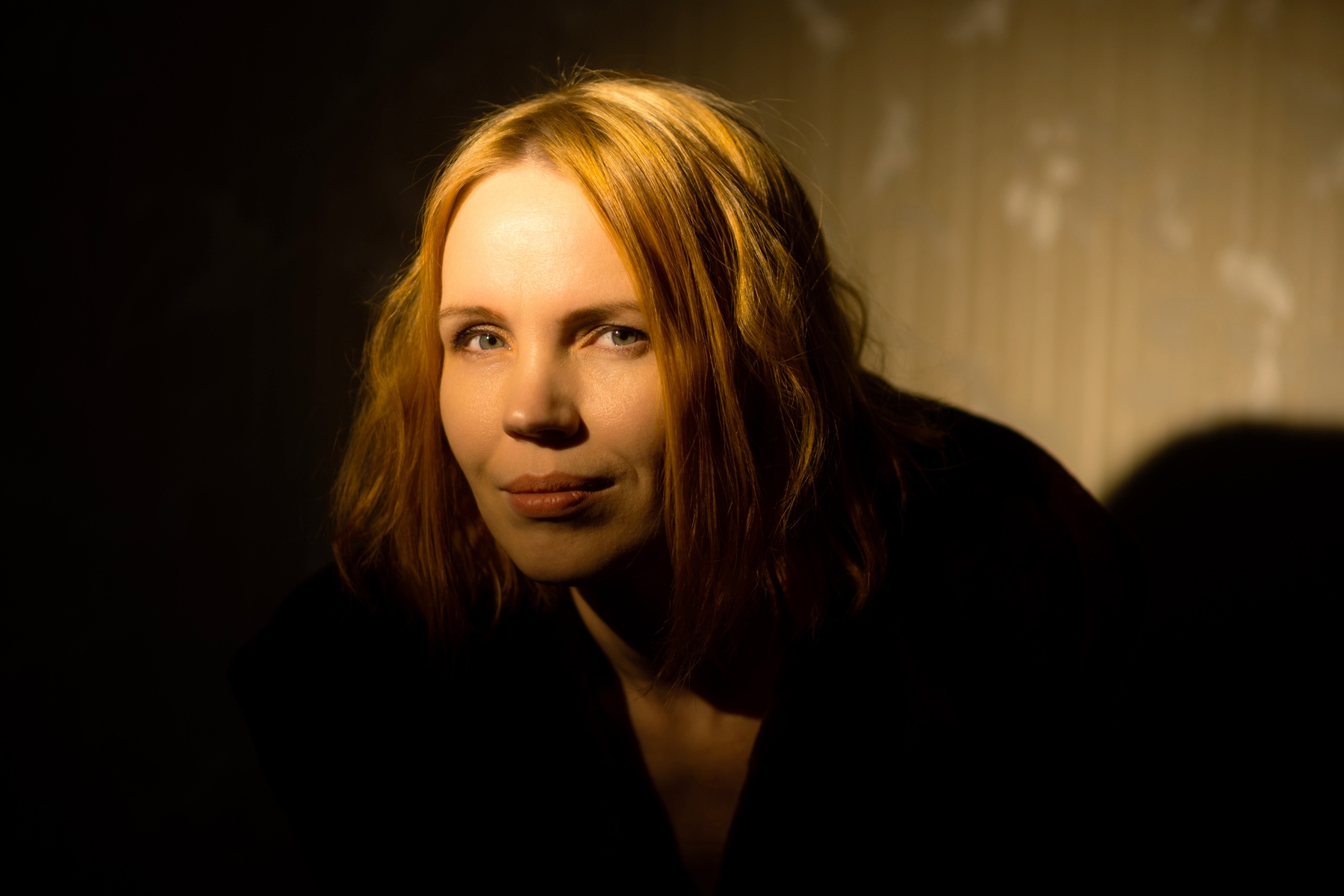 A woman with shoulder-length blonde hair is partially illuminated against a dark background. She is wearing a black top and has a neutral expression. The lighting creates a dramatic effect with shadows on one side of her face.