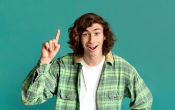 A person with long, wavy brown hair wearing a green plaid shirt and a white t-shirt underneath. They are smiling and raising their index finger as if having a great idea. The background is a solid teal color.