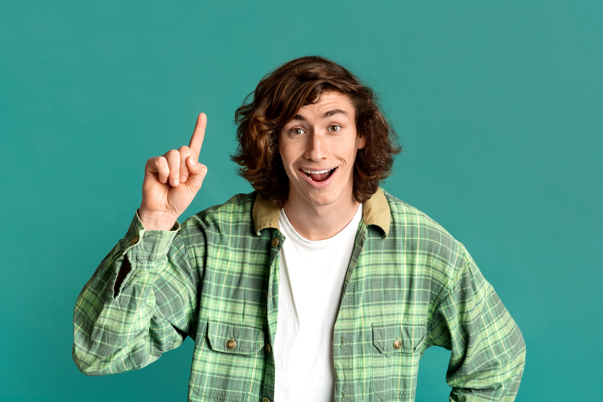 A person with long, wavy brown hair wearing a green plaid shirt and a white t-shirt underneath. They are smiling and raising their index finger as if having a great idea. The background is a solid teal color.